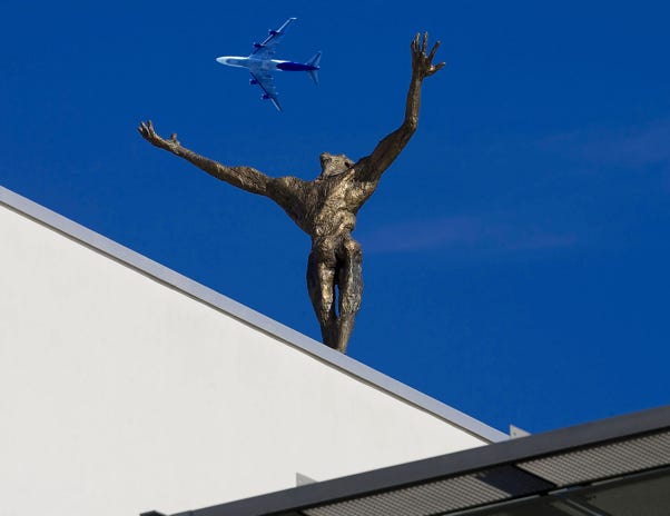 Statue auf Mainzer Staatstheater streckt die Hönde gen Himmel. Dazwischen fliegt ein Flugzeug.